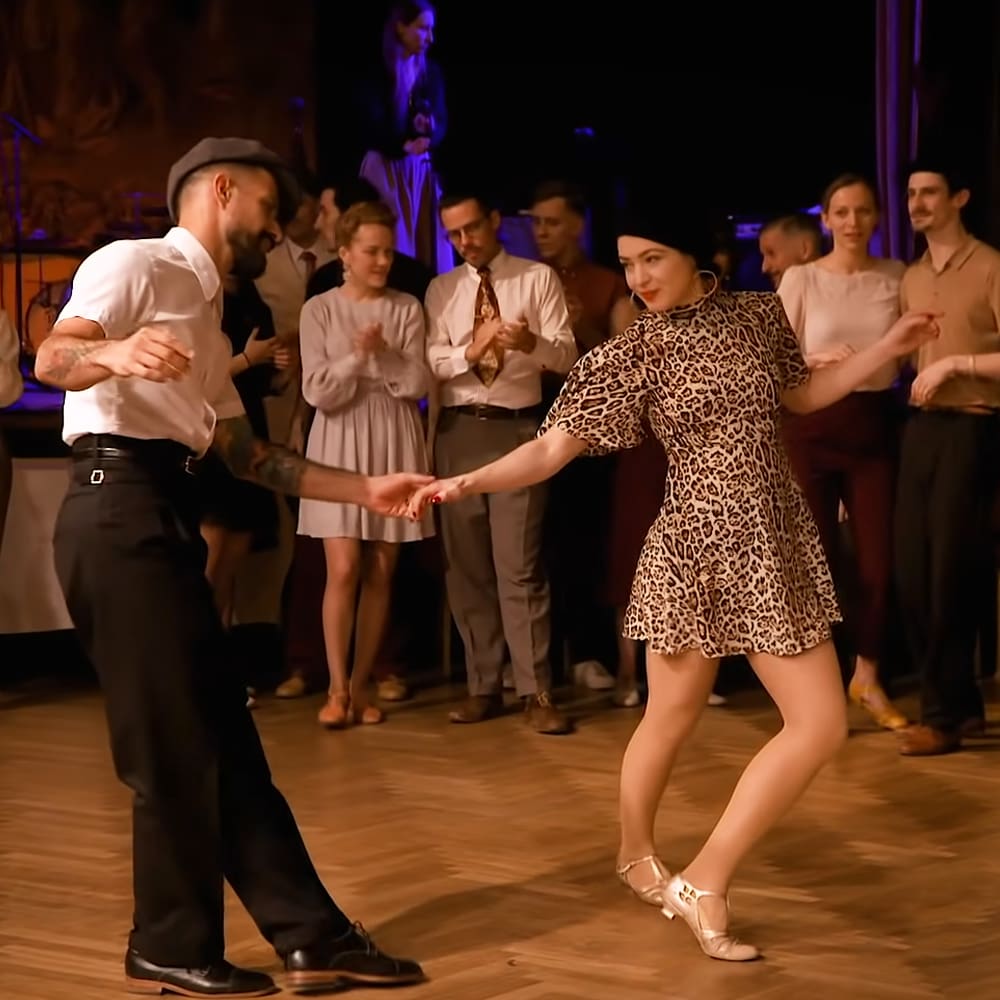 Adult male and female dancers in a social dance setting, possibly a ballroom or event hall; man wears a white shirt with black trousers and dark shoes, woman sports a leopard print dress and ballet flats