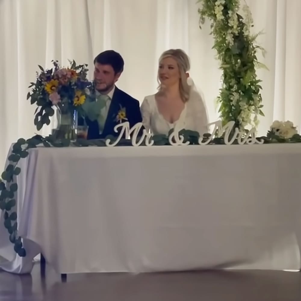 A wedding couple at their reception table in an indoor venue with a white curtain backdrop, decorated with floral arrangements including sunflowers.
