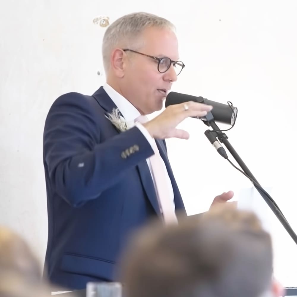 A middle-aged man giving a speech at an event, possibly a wedding or formal gathering, indoors against a plain white wall with bright diffused lighting.