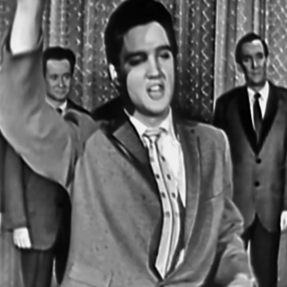 Young adult male in a light-colored suit jacket with pinstripes stands at the forefront, surrounded by other adults in formal attire against an indoor stage backdrop.