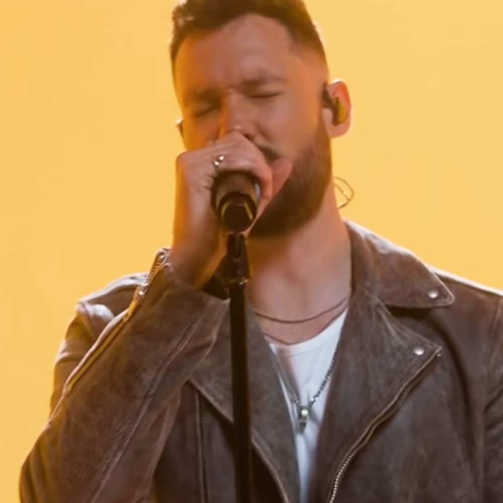 An adult male performer on stage, wearing a dark brown leather jacket with zipper and snap buttons, white t-shirt underneath, thin necklace or chain, in an indoor venue with solid yellow backdrop from stage lighting.