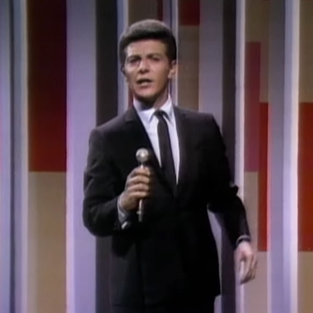 A young adult male in a dark suit jacket and trousers performing on stage in what appears to be a television studio with vertical striped backdrop.