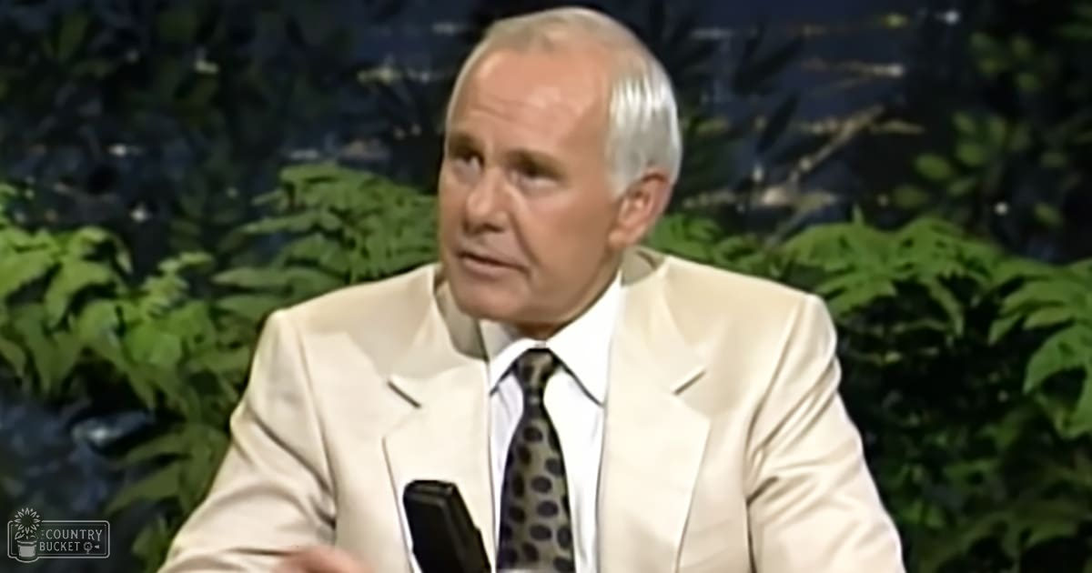 An older adult male singer wearing a light-colored suit jacket, white dress shirt, and dark tie with lighter dots or circles, standing in front of a microphone in a studio setting with artificial greenery.
