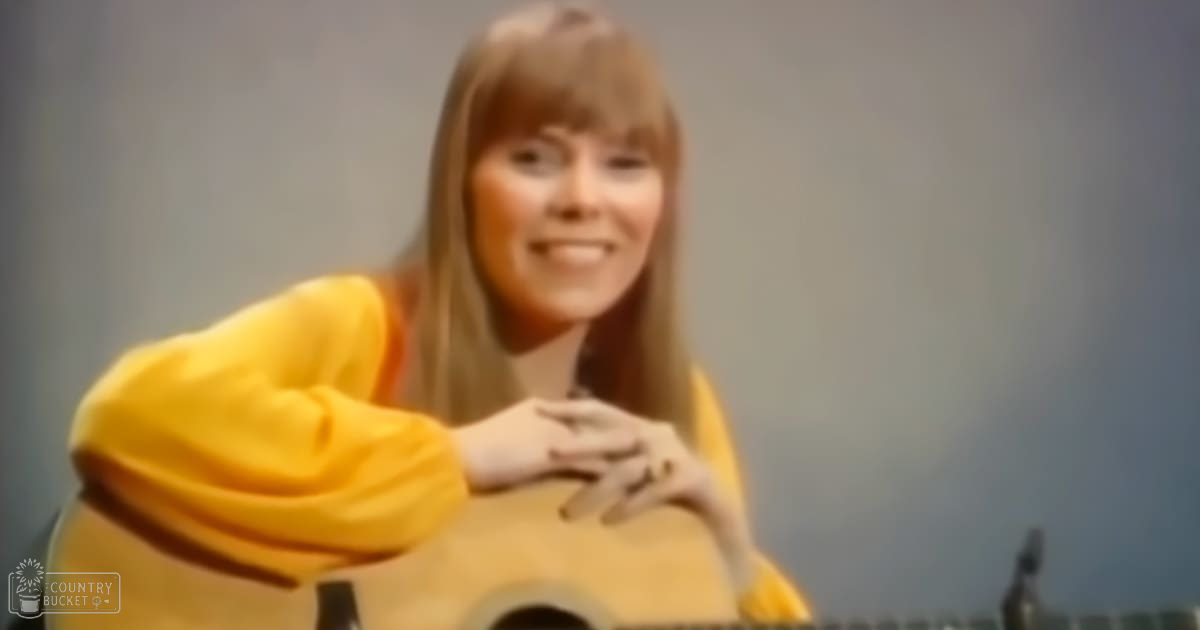 A young adult female wearing a bright yellow, loose-fitting long-sleeved garment indoors with a guitar partially visible under her arms.