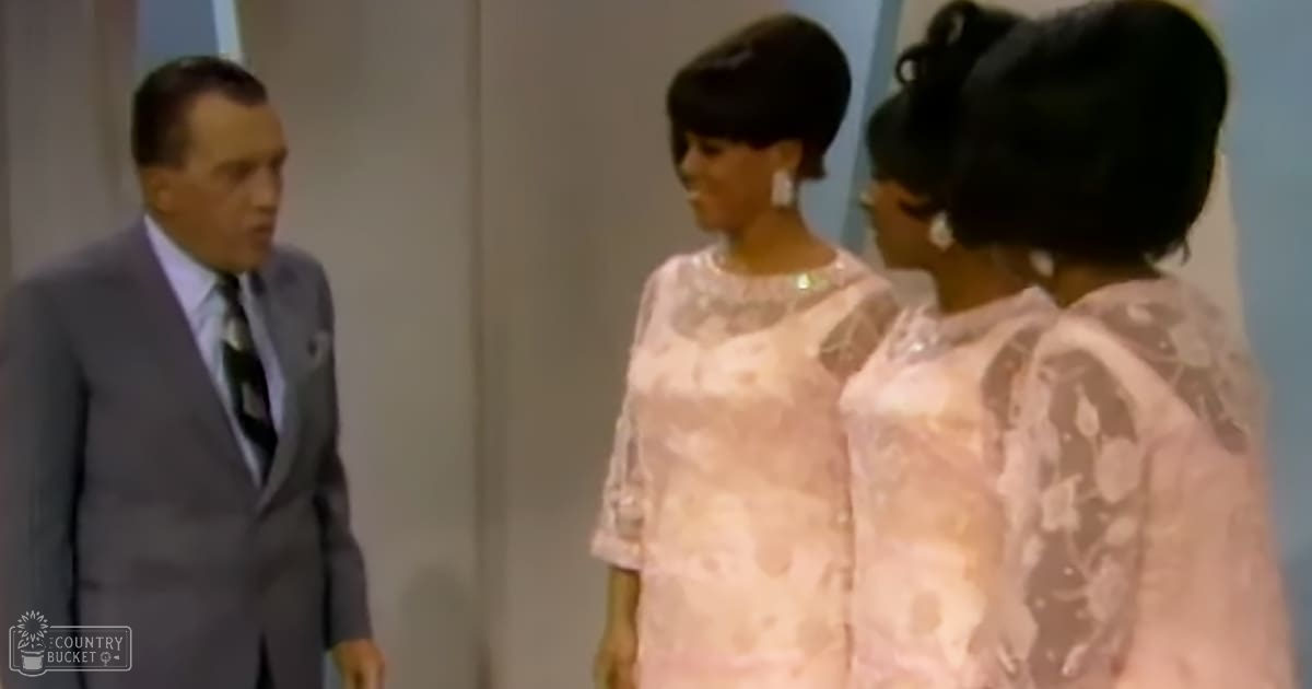 An adult male wearing a well-fitted gray suit with a white shirt and dark tie stands indoors, surrounded by two women dressed in light pink floral dresses, indicating a formal or professional setting.