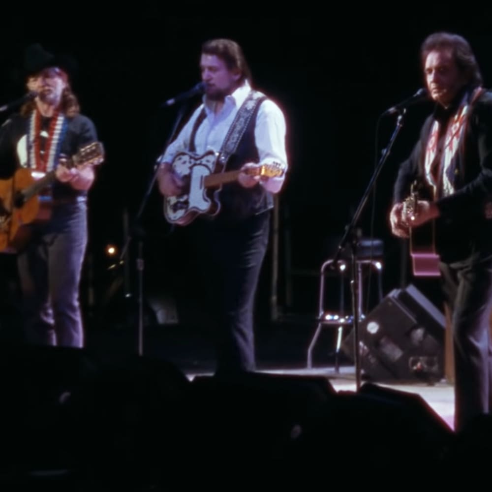 An adult male singer in a dark-colored shirt and red necklace performs alongside other musicians at an indoor concert venue under dramatic stage lighting.