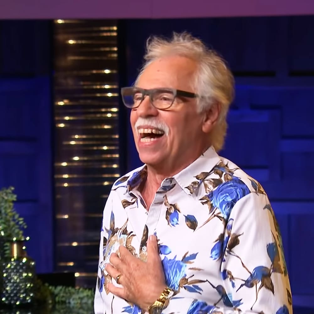 An older adult male in a casual, floral print shirt with rectangular glasses and a gold watch, set against dark blue studio walls.