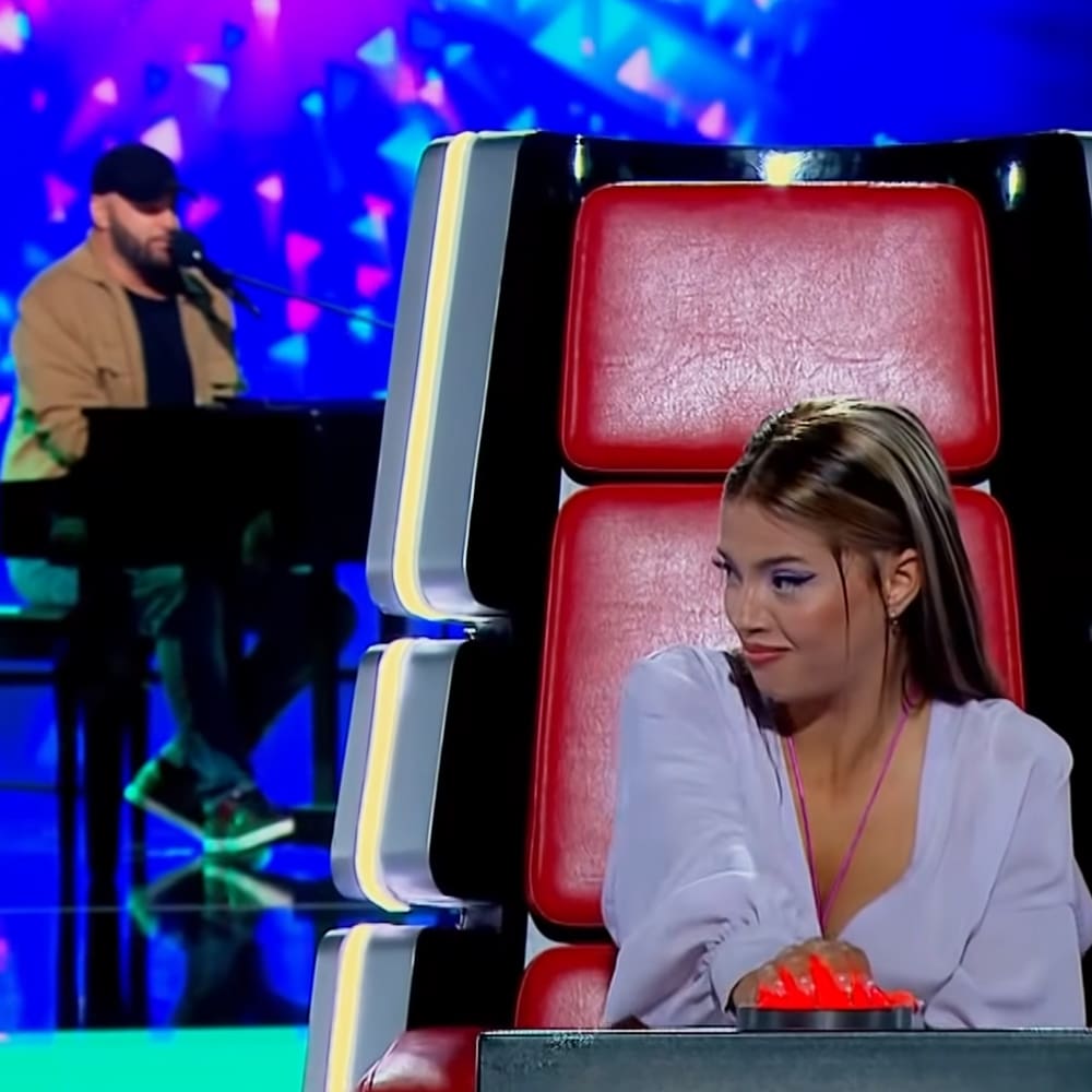 A young adult female in a talent show or singing competition setting, wearing a white blouse with accessories and seated on a red chair.