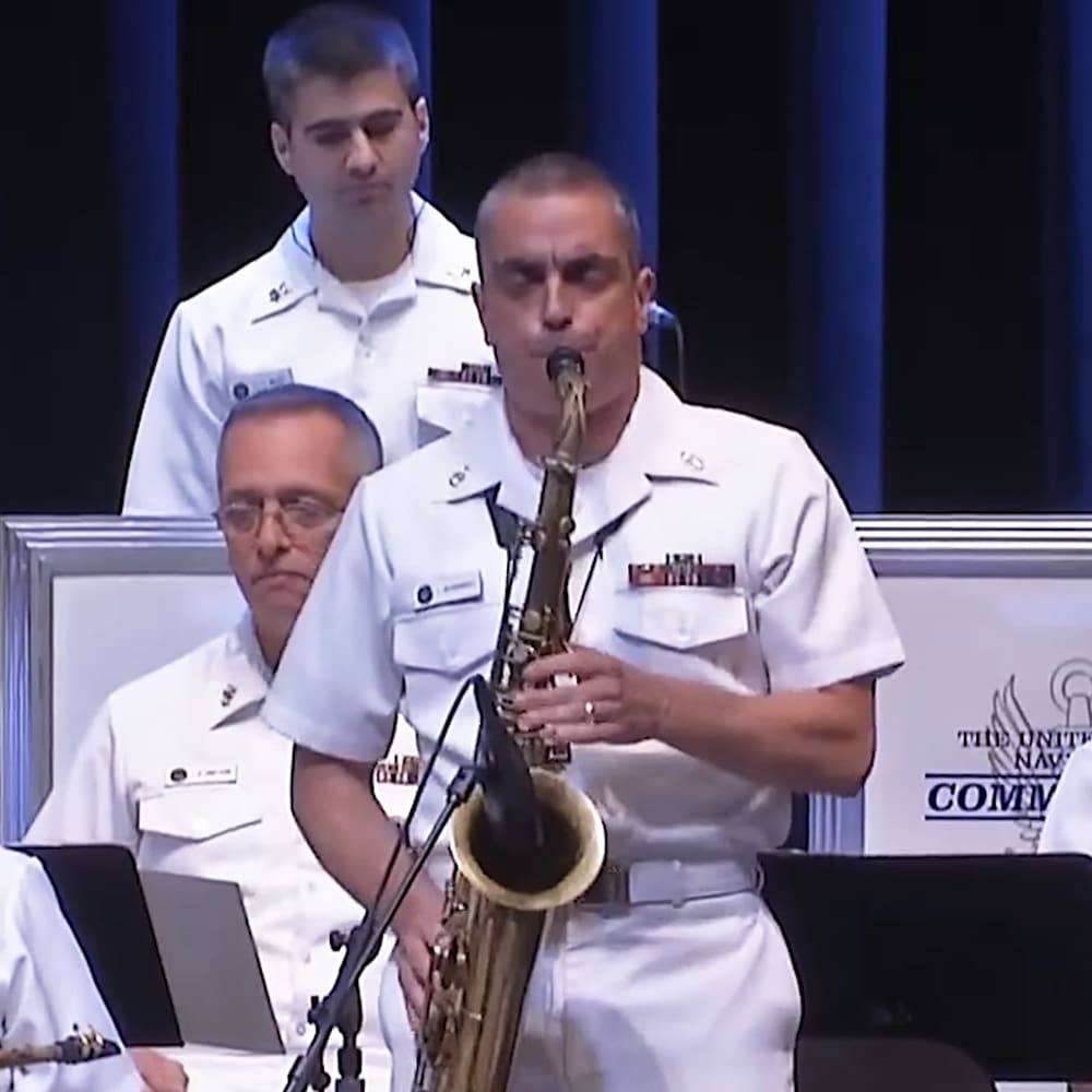 A man playing a saxophone in front of a backdrop with 'The United States Navy' written on it, at an indoor event hosted by the U.S. Navy.