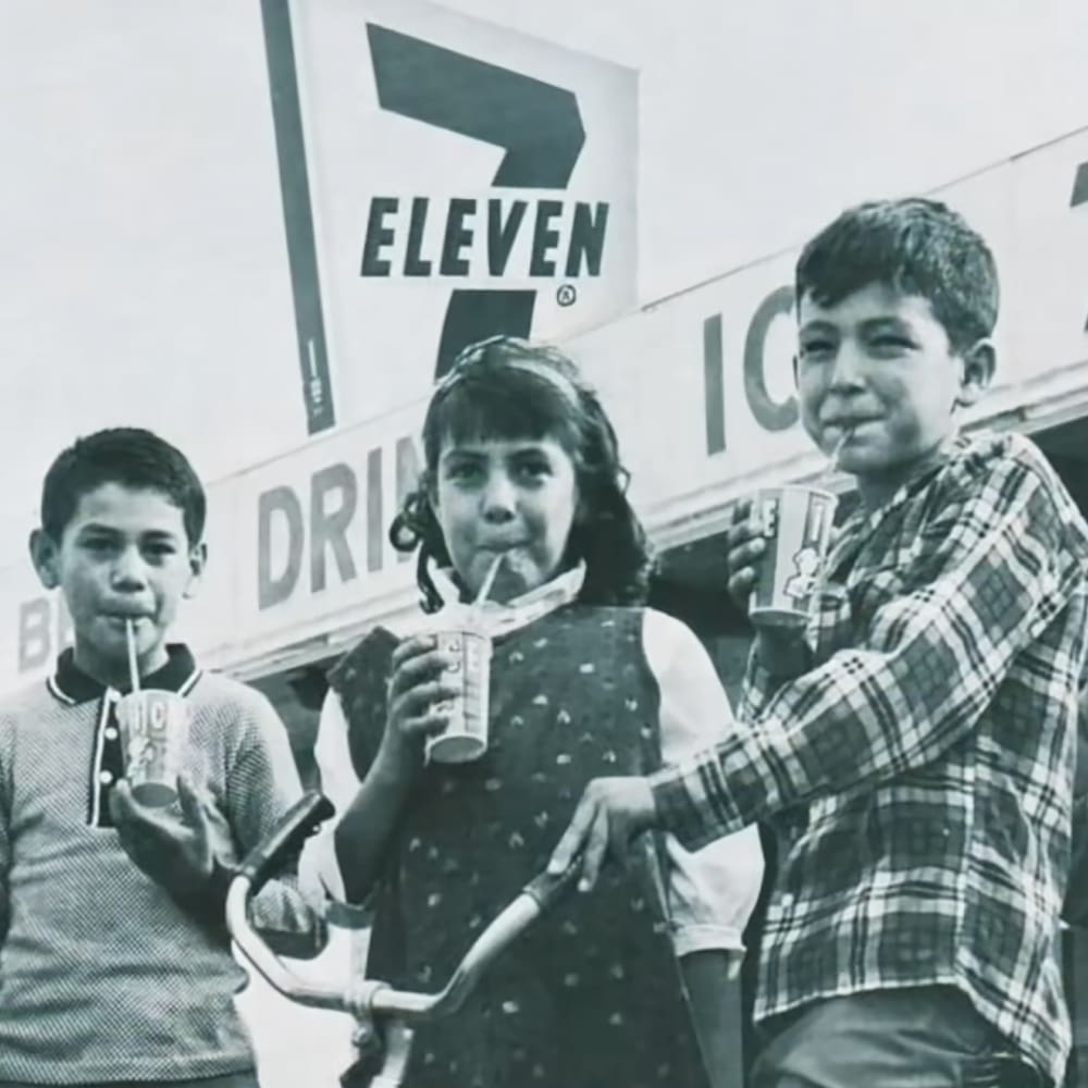 Group of children in front of a 7-Eleven store with vintage clothing from the mid-20th century, holding drinks and a bicycle handlebar.