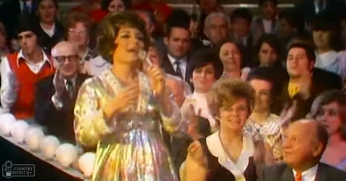 A young to middle-aged female performer wearing a floral-patterned dress in pastel colors and glasses stands on stage, with an audience of adults seated behind her. The setting appears to be a theater or concert hall during daytime.
