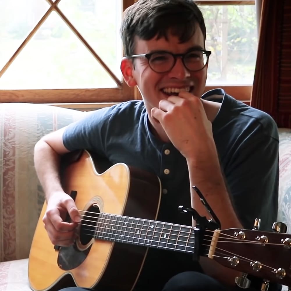 A young adult male wearing a dark-colored, relaxed fit short-sleeved shirt with glasses plays guitar in an indoor setting with natural light.