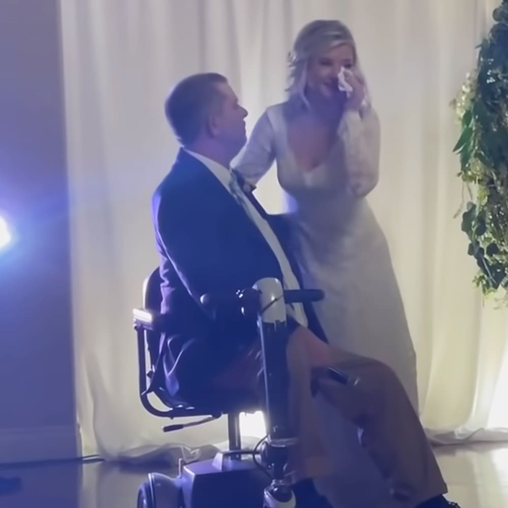 A wedding scene with an adult male singer wearing a dark blue suit with white shirt and light-colored tie, seated in a wheelchair; a female figure stands beside him in a white dress with long sleeves. The setting includes white curtains and stage lighting.