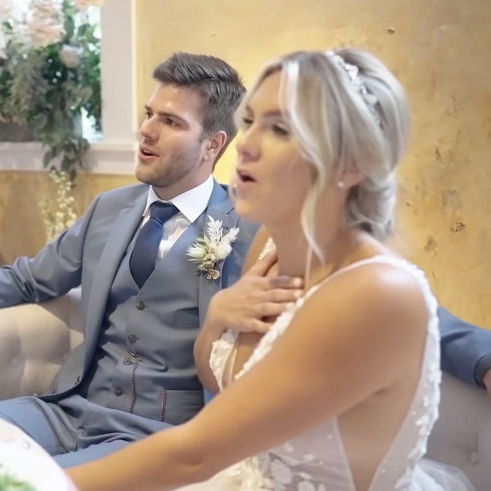 A couple at a wedding ceremony indoors with a young adult male wearing a light gray suit and white dress shirt, and an adult female in a white wedding gown holding her hand over her chest. The background includes floral decorations and greenery.