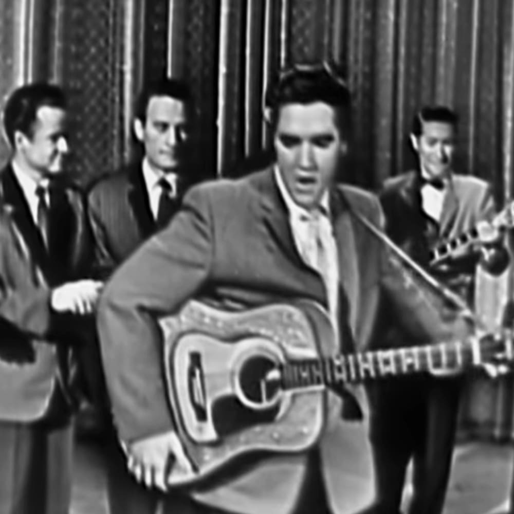 A mid-20th-century performance scene with an adult male performer in a suit jacket and tie, holding a guitar labeled 'MARTHA & THE', surrounded by other similarly dressed individuals on stage under artificial lighting.