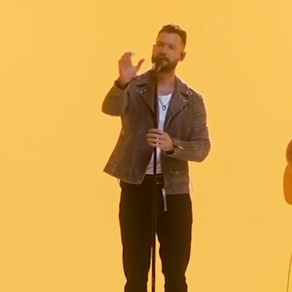 An adult male singer in a dark brown leather jacket over a white t-shirt with accessories, performing on stage under bright yellow lighting.