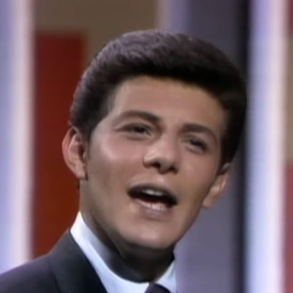 Young adult male in a mid-20th century formal suit, standing against a colorful stage backdrop in an indoor studio setting.