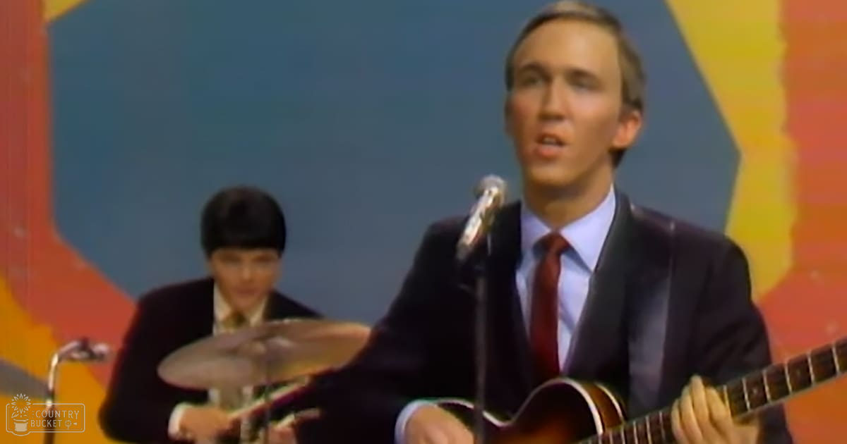 A young adult male singer wearing a dark suit jacket with classic fit, white dress shirt, and red tie is playing guitar while singing into a microphone. A teen to young adult male drummer in a dark blazer and clean collared shirt plays drums behind him on a stage with vibrant geometric shapes.