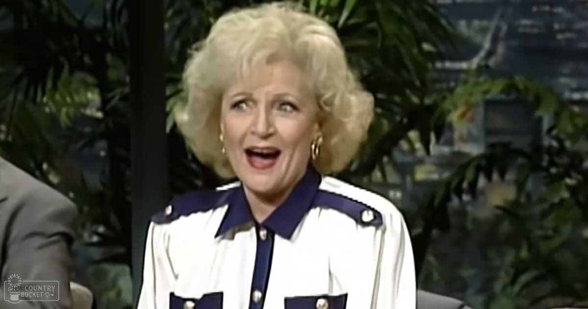Older adult woman wearing a white and navy blue patterned jacket with gold hoop earrings, set against an indoor studio backdrop with plants and a nighttime urban scene.
