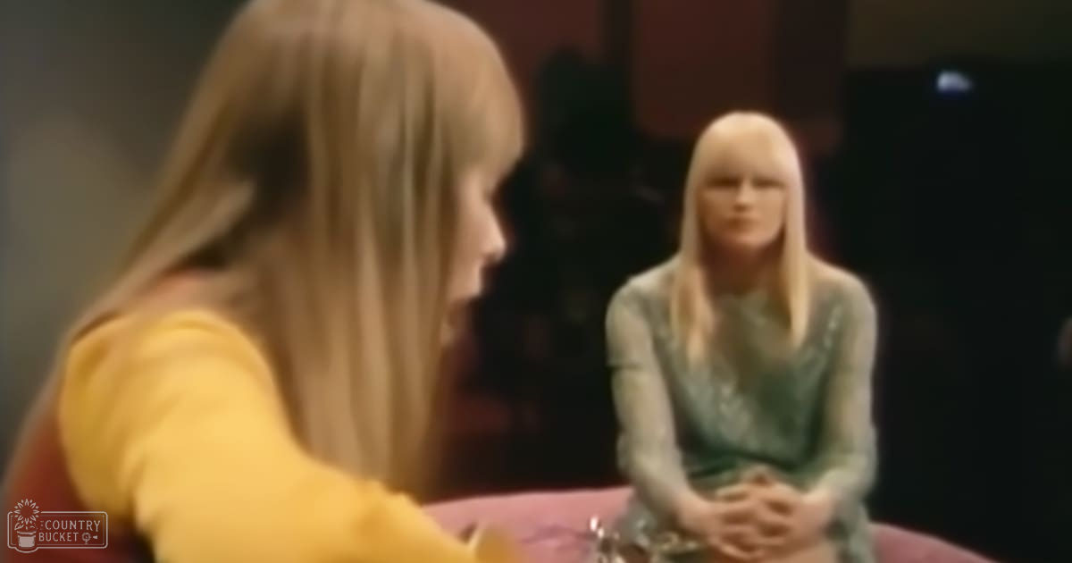 Two women are seated indoors under artificial lighting; one wears a yellow top with short sleeves and cotton-like material from the late 20th century, while the other dons a green, patterned dress in similar style.