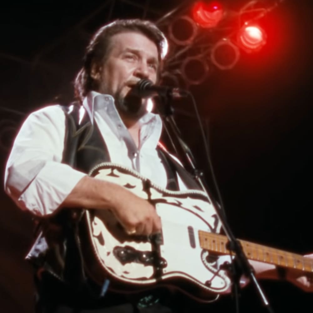 Middle-aged male singer in white button-down shirt with rolled-up sleeves and dark vest, performing at a concert venue with red stage lighting.