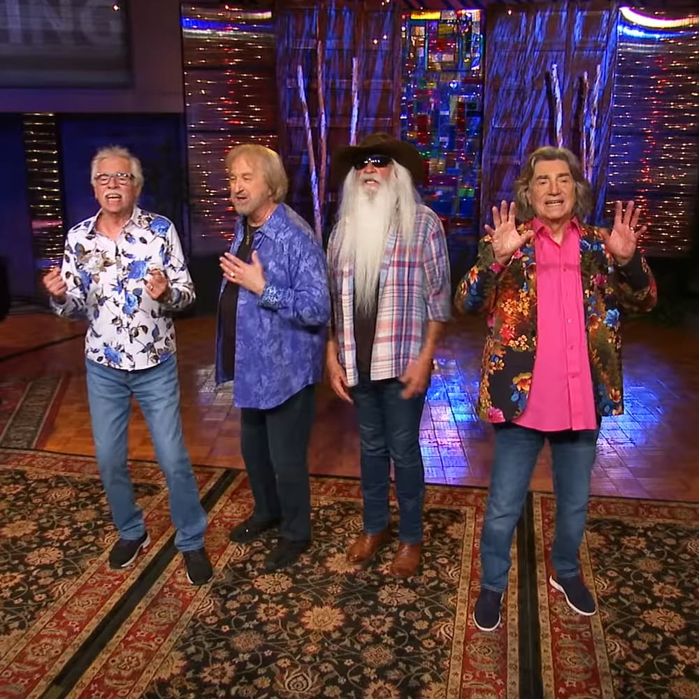 An older adult male singer in a white shirt and blue floral patterns on stage during a talk show or variety program performance, surrounded by other performers under colorful artificial lighting with stained glass windows and reflective curtains in the background.