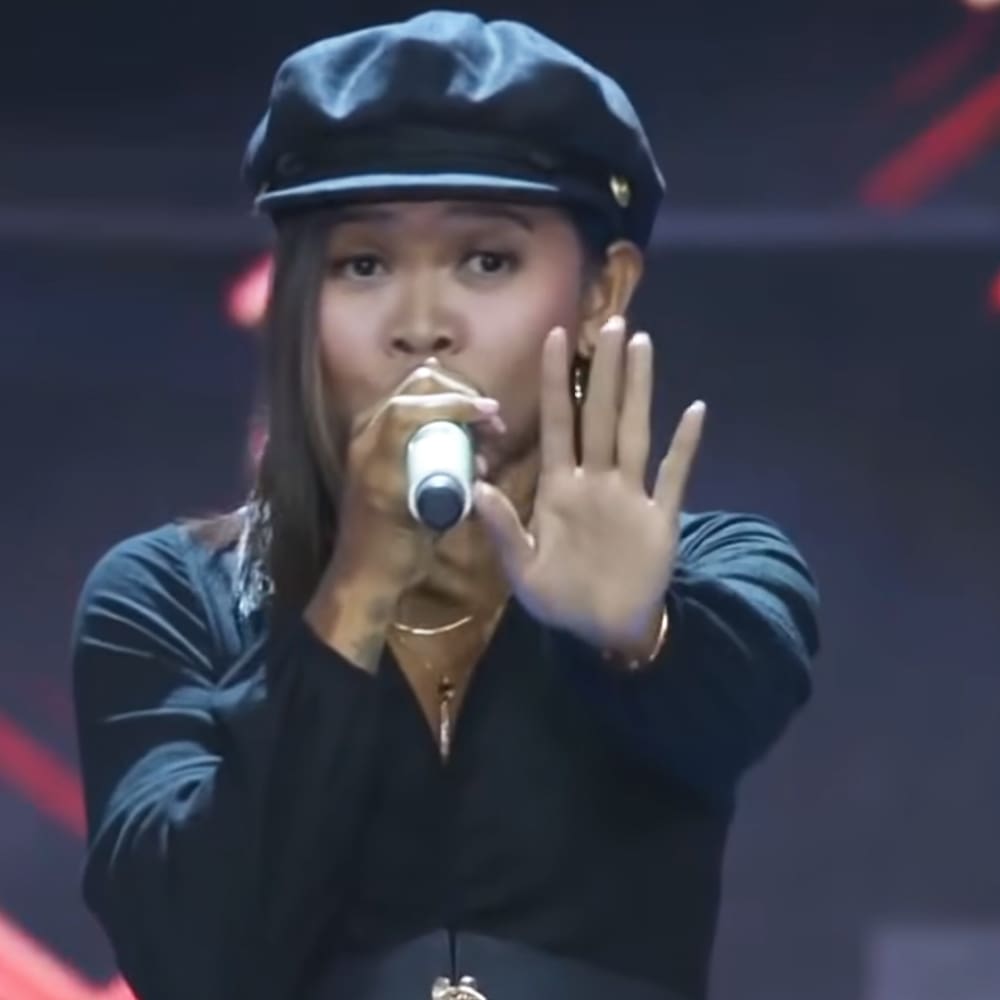 Young adult female singer in a relaxed, loose-fitting long-sleeved dark blue or black top with low neckline and gold necklace performing on stage with red lighting accents.