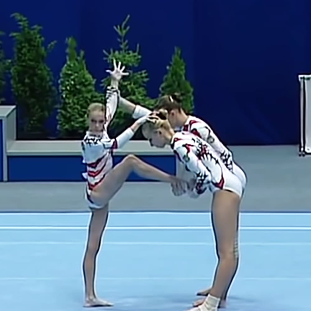 Young adult female gymnasts performing in an indoor competition venue with blue walls and artificial trees, wearing white leotards with red and black stripes under artificial lighting.