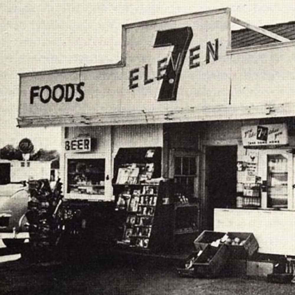 Urban 7-Eleven store with 'FOODS' sign, vintage car, and shelves stocked inside the building during daytime in an urban setting