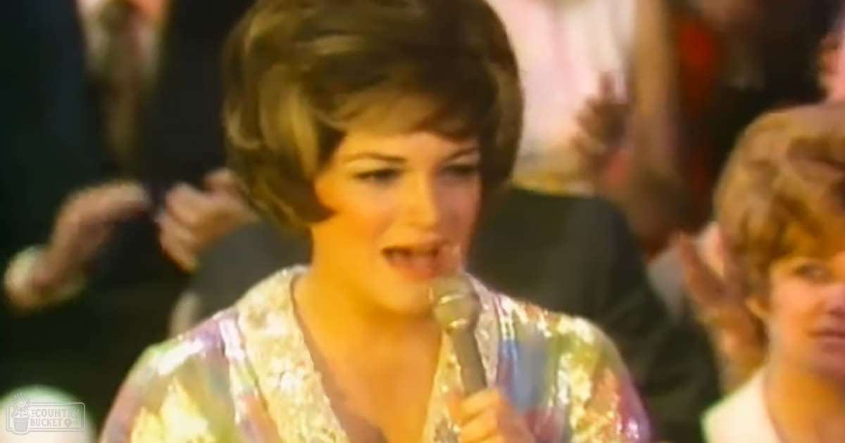 A young adult female sings into a microphone at an indoor event under warm artificial lighting, with blurred background figures in formal attire.