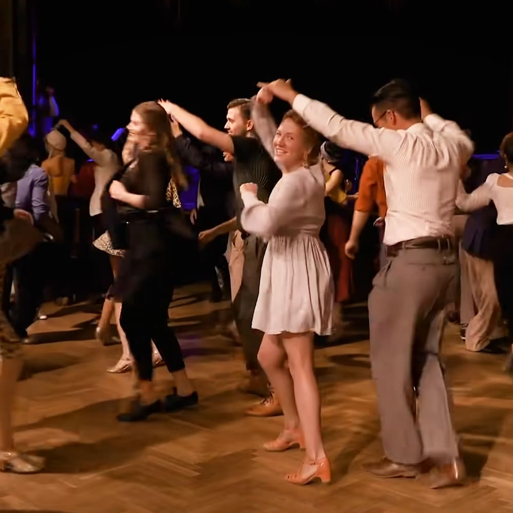 A dance floor scene at an indoor event with a young adult female wearing a white knee-length dress and middle-aged man in light gray trousers, set against wooden flooring typical of ballrooms under artificial lighting.