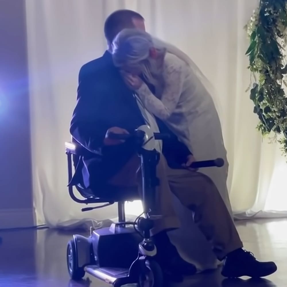 Adult male seated in a motorized wheelchair at an indoor event with white curtains and floral decor, bright lighting casting shadows.
