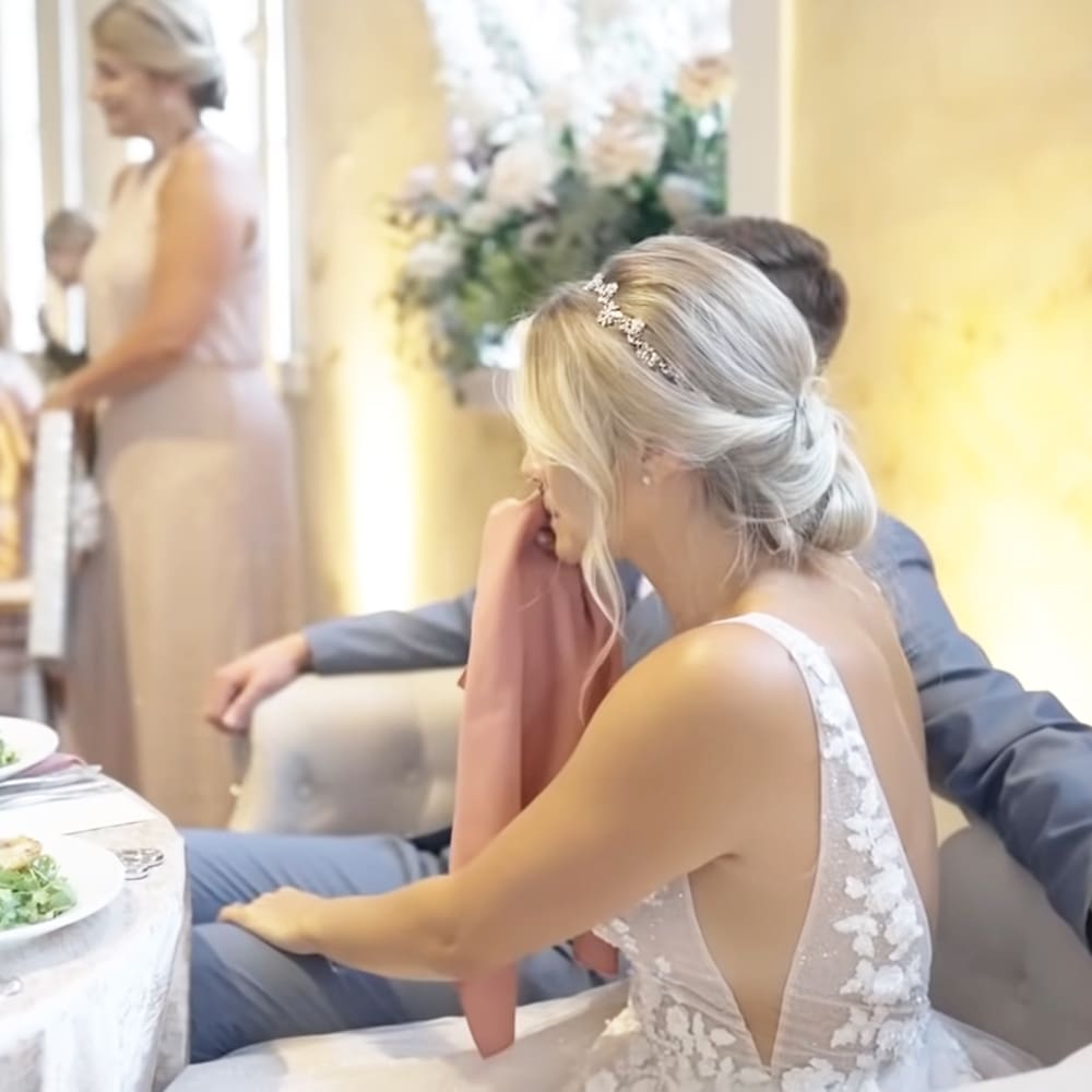 A bride in a white lace wedding dress at her reception with other guests and a table setting, likely indoors during the day.
