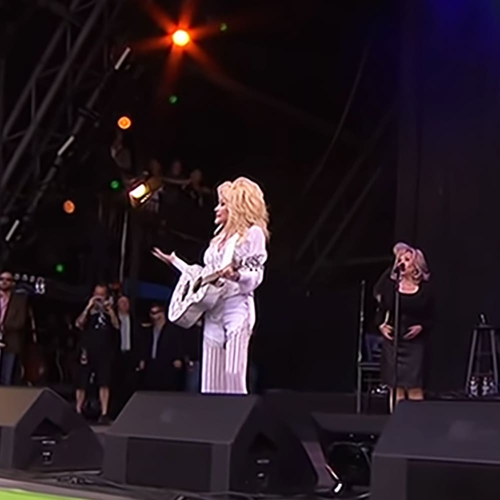 Adult female on stage at an outdoor concert, wearing a white and black striped outfit with microphone in hand; background includes another adult female performer and stage lighting creating starburst effects.