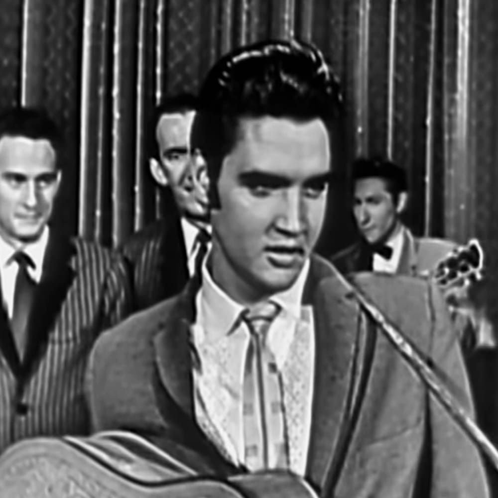 A young adult male singer wearing a patterned tie, seated on stage with a microphone and guitar, performing alongside other musicians dressed in formal attire.