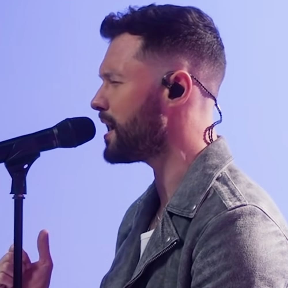 A male adult in his 30s-40s wearing a denim jacket over a white shirt performs on stage with a microphone stand and earpiece visible.