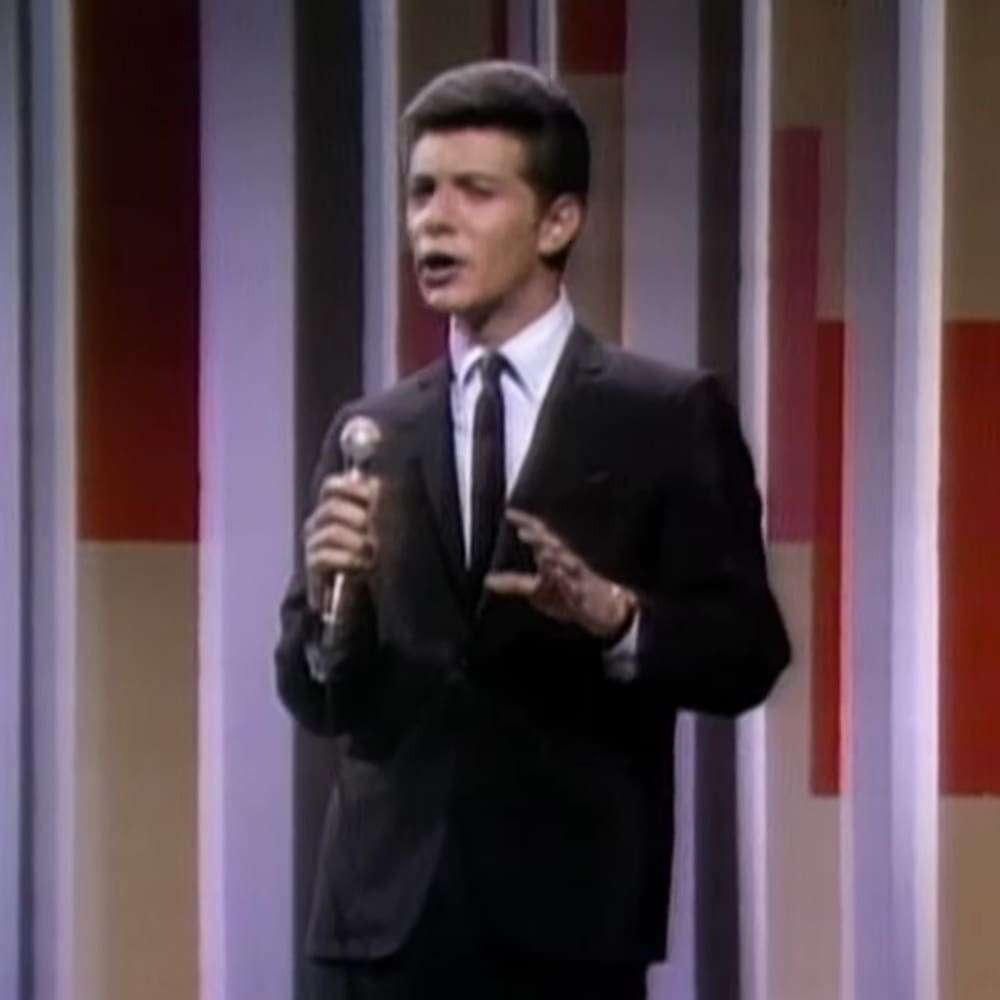 A young adult male in a formal suit jacket and trousers, performing on stage with a microphone against a striped backdrop.