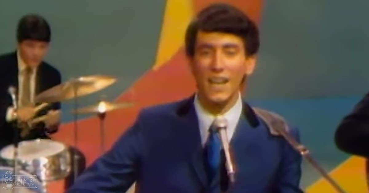 A young adult male singer performs into a microphone, dressed in a mid-20th-century dark blue suit with white shirt and blue tie; a drummer in the background wears a patterned tie and holds drumsticks.