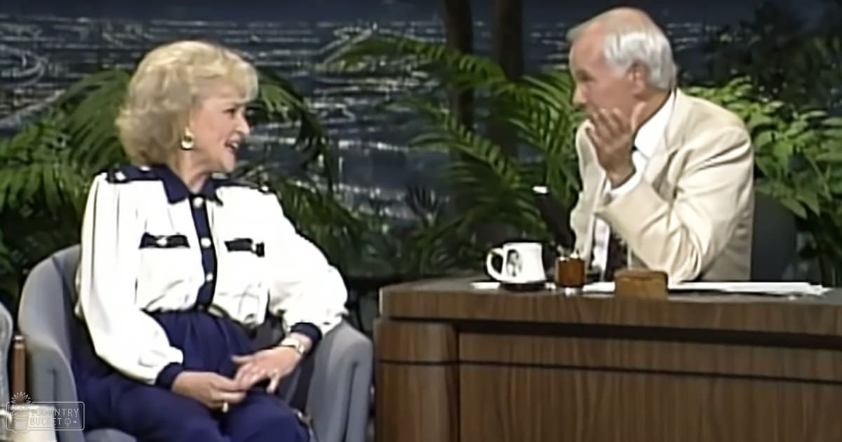 Two adults engaged in conversation in a talk show setting with decorative plants, city lights at night, and a round wooden table.