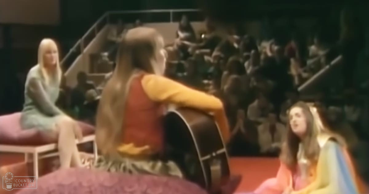 A woman with long brown hair plays an acoustic guitar, surrounded by other performers on red cushioned benches in a darkened room during what appears to be a musical performance or concert.