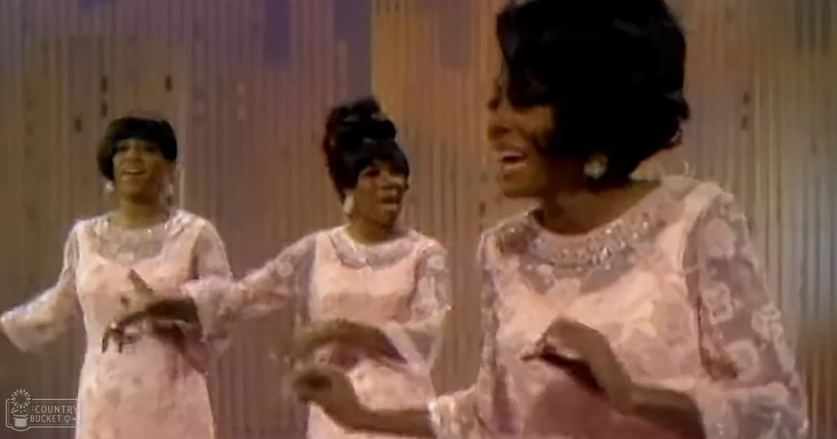 Three women performing in a studio setting, wearing light pink dresses with floral patterns from the 1960s or early 1970s; vertical wooden paneling suggests an indoor stage environment.