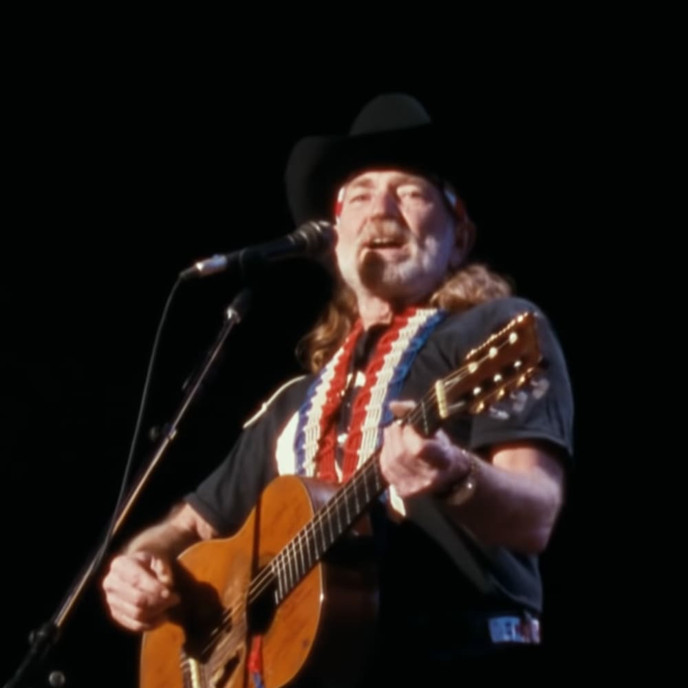 Middle-aged male performer wearing a black cowboy hat, dark-colored t-shirt with red, white, and blue pattern, long curly hair, and watch; performing at an indoor concert stage under focused lighting.