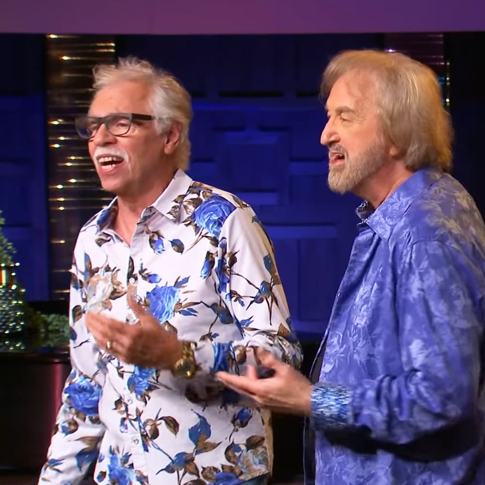 Two middle-aged men in a studio setting, wearing floral shirts and blue patterned shirts with relaxed fits and button-up collars. Dark blue walls and decorative elements suggest an interview or talk show environment.