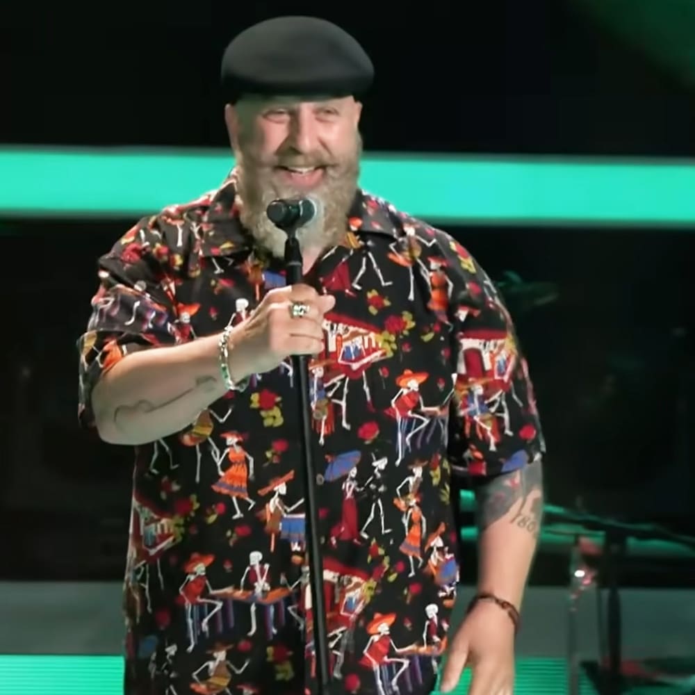 A middle-aged adult male singer wearing a patterned black short-sleeved shirt with colorful human figures in red, blue, yellow, and white, paired with a black beret-style hat and multiple bracelets. Microphone stand with microphone suggests an event or performance setting.