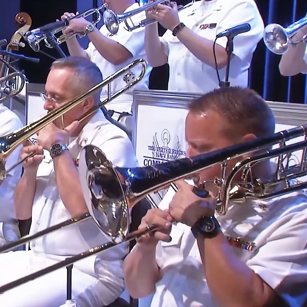 A group performing with trombones in an indoor setting, likely a concert hall. White uniforms dominate the scene against a backdrop reading 'The United States Navy Band.'