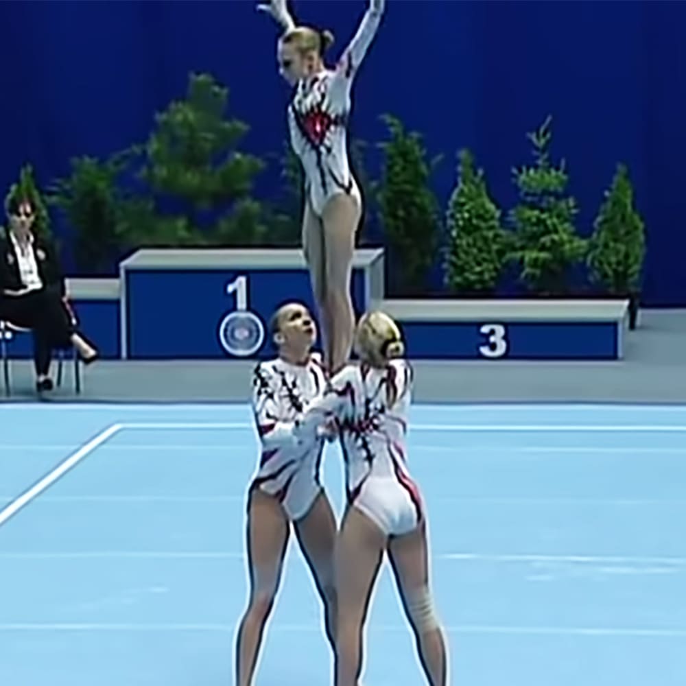 A group of young adult female gymnasts perform in a competition setting, wearing white leotards with black and red patterns against a blue wall with numbered sections.