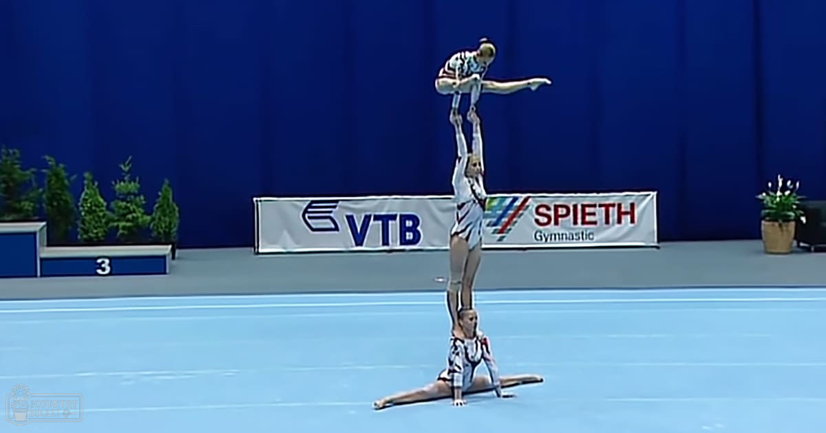 A gymnastics performance by two young adult female gymnasts wearing matching white and black leotards with a pattern of small dark shapes, barefoot on an indoor blue gymnastics mat with banners in the background.