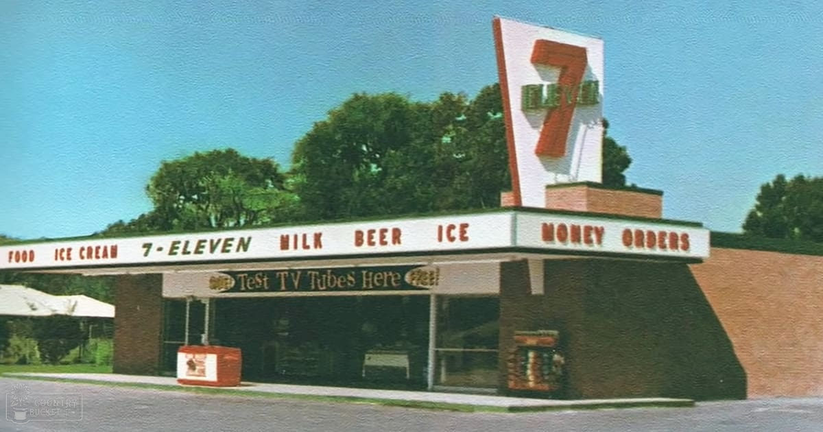 An outdoor 7-Eleven convenience store sign with '7-ELEVEN' prominently displayed, red and white awning over the entrance, trees in background, clear blue sky, mid-20th century era, daytime bright sunlight