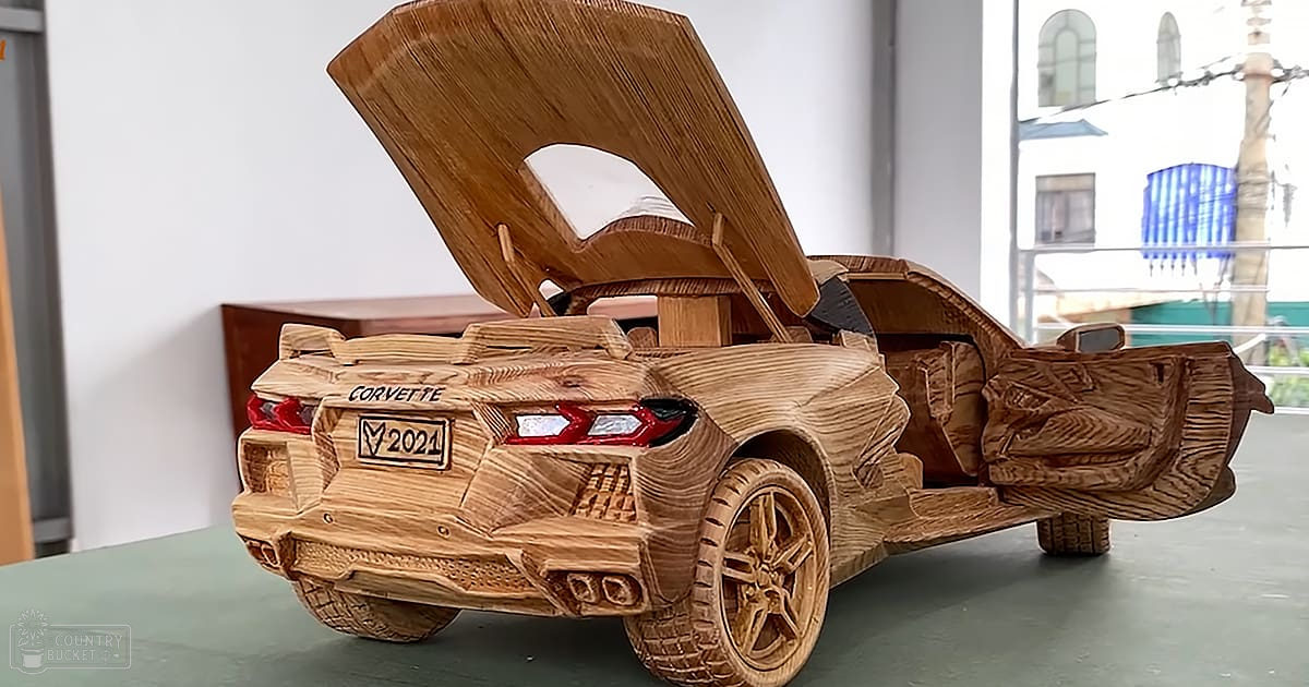 Detailed wooden model of a Chevrolet Corvette 2021 in an indoor art gallery setting; detailed textures, open doors revealing interior, visible windows showing outdoor elements.