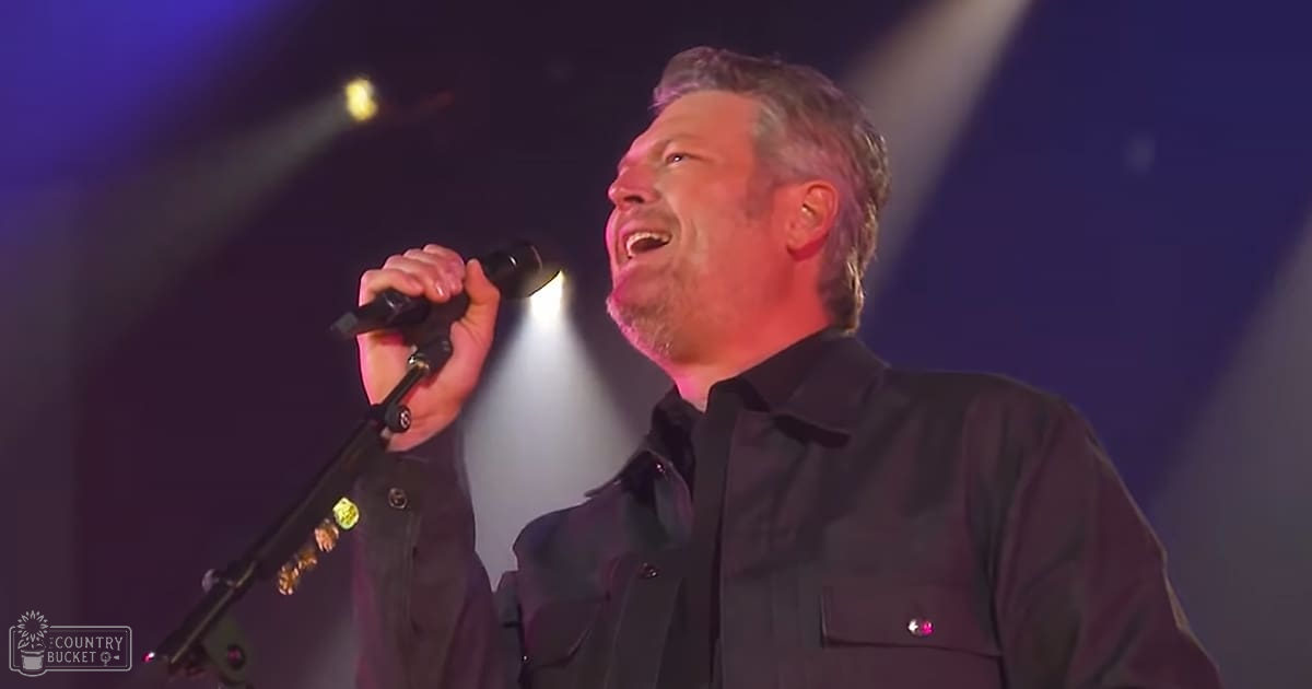 A middle-aged male performer in a relaxed black button-down shirt with pockets, standing on stage under dramatic lighting during an evening event.